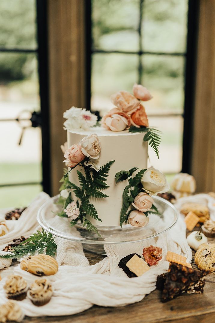 Single tier white iced wedding cake with roses and foliage surrounded by cupcakes and cookies 