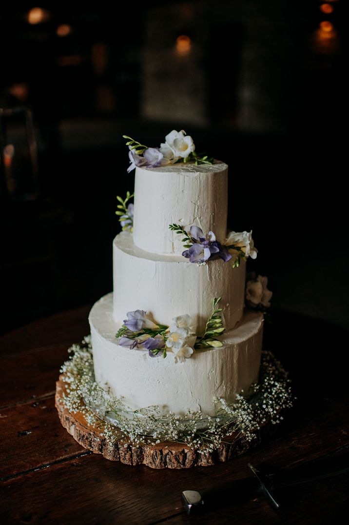 Rustic barn wedding with a three tier frosted white wedding cake with purple and white flowers on tree trunk stand 