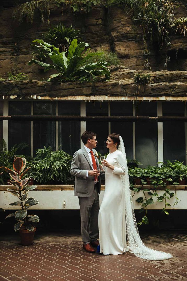 Groom in grey suit and bride in long sleeve satin wedding dress standing in The Barbican Conservatory glasshouse wedding venue 