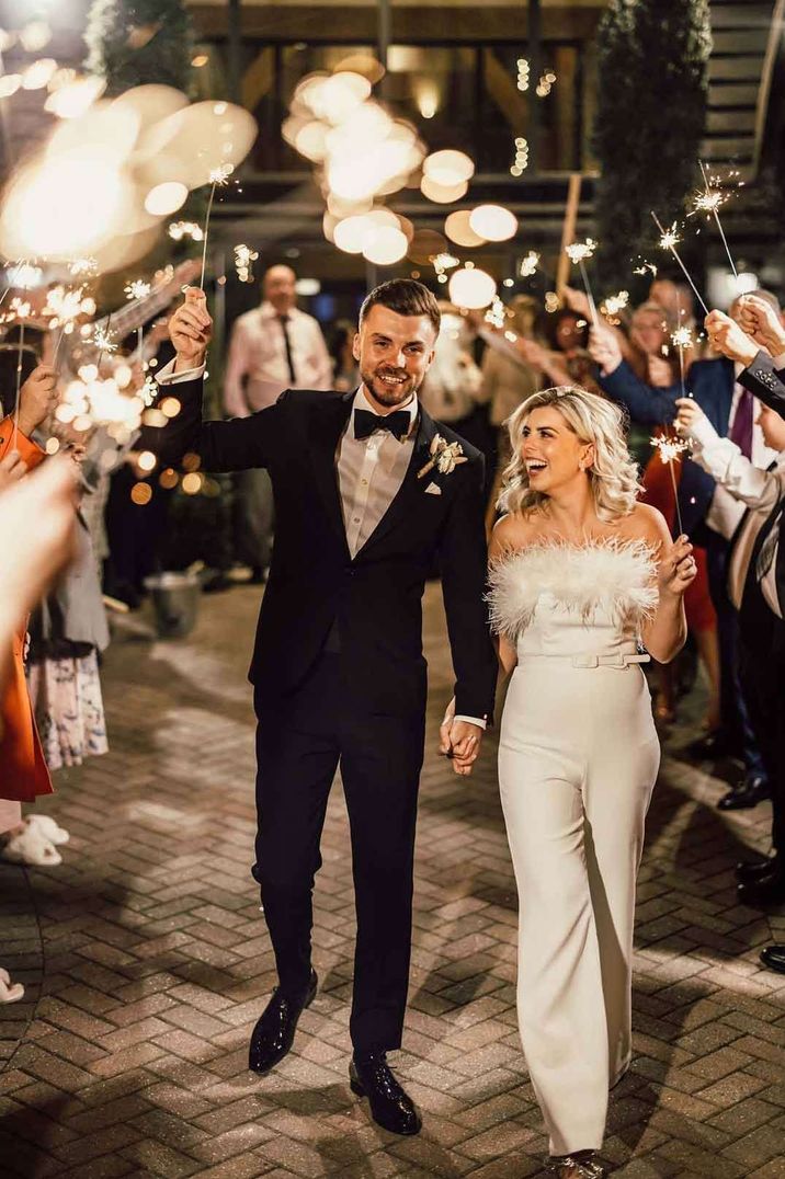 Bride in strapless wedding feather jumpsuit walking with groom in classic black tuxedo doing sparkler exit 