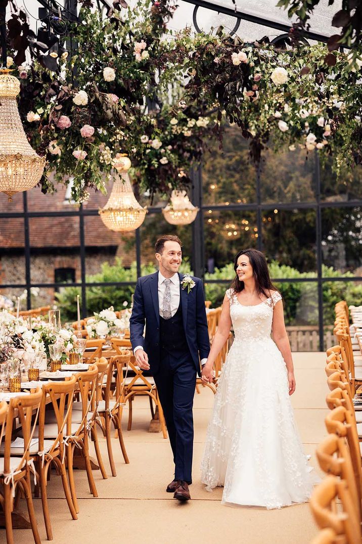 At home glass marquee with chandeliers and hanging large floral installation with the bride in floral applique wedding dress with the groom in navy suit 