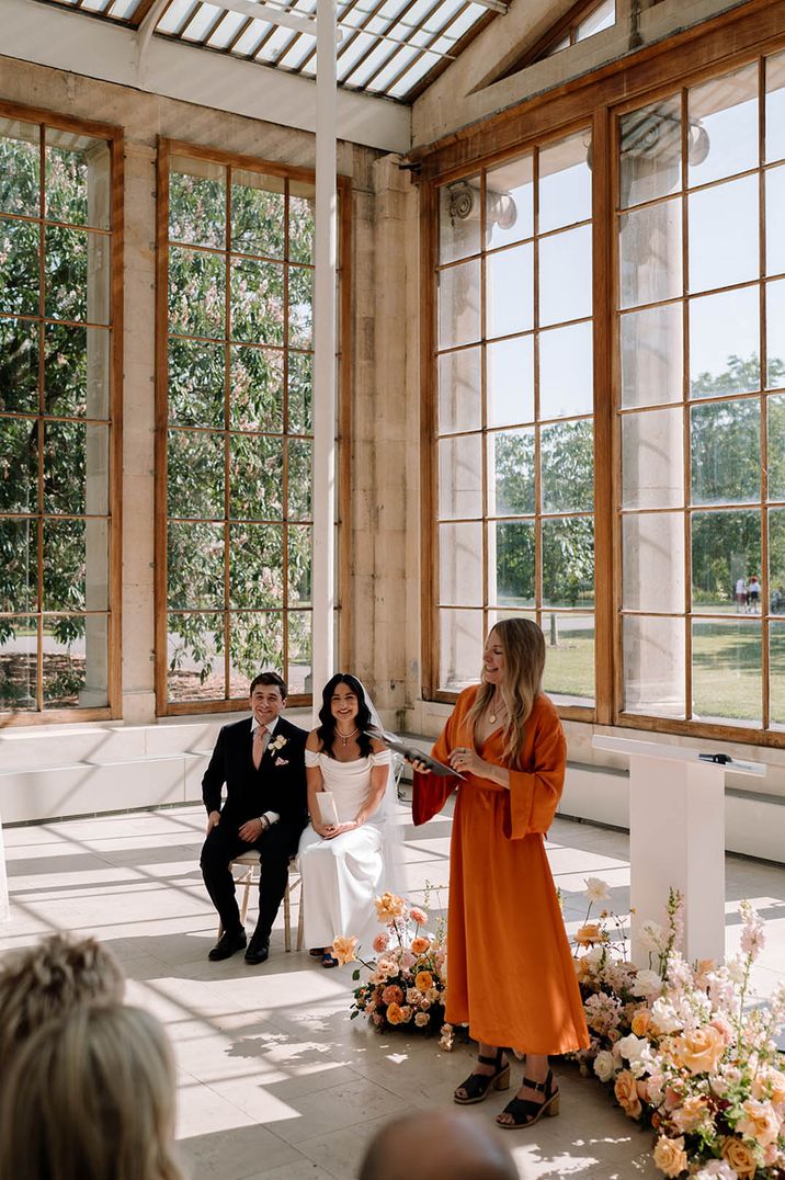 Roxy Celebrates Love in orange jumpsuit at wedding ceremony with bride and groom watching from chairs 