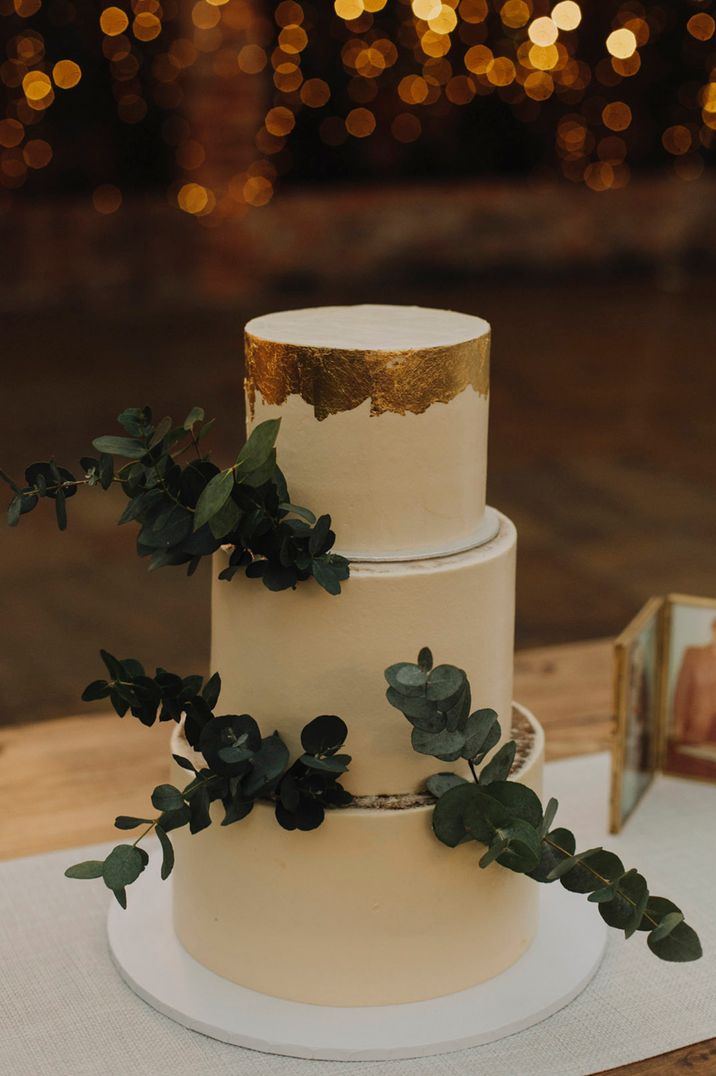 Three tier white wedding cake topped with gold and decorated with greenery 