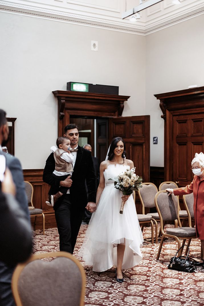 Bride in tulle strapless gown with high low wedding skirt walking back down the aisle with the groom and their toddler 