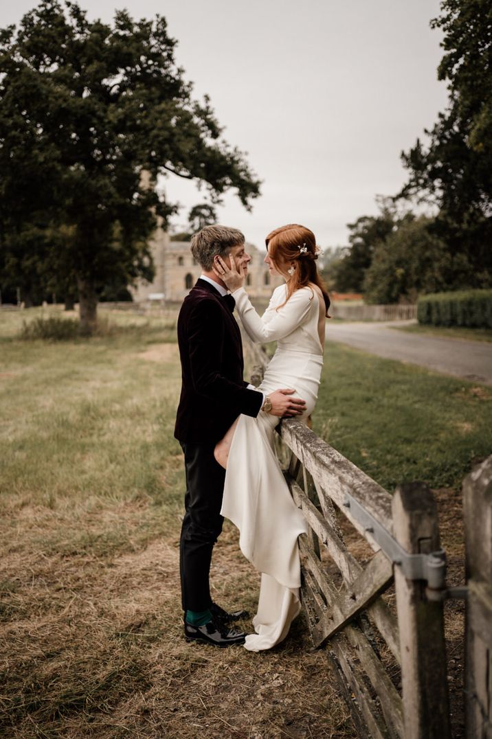 Bride in a minimalist wedding dress with plain long sleeves sitting on a fence embracing her husbands face