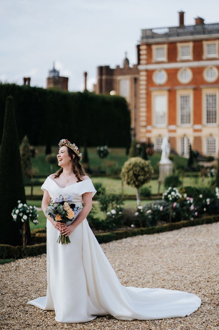 Bride in an off the shoulder wedding dress and flower crown standing in the gardens at Hampton Court Palace