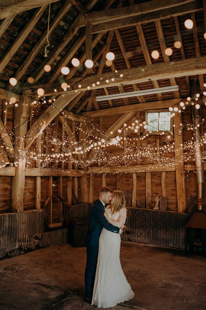 Bride in short sleeved embellished wedding dress and groom in dark suit dancing in a barn covered in fairy lights