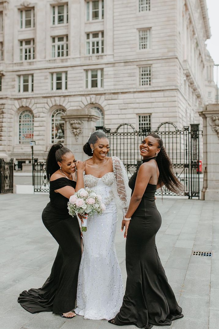 Bride wears fitted lace wedding dress and stands in the middle of her bridesmaids in black bridesmaid dresses in different styles for black tie wedding