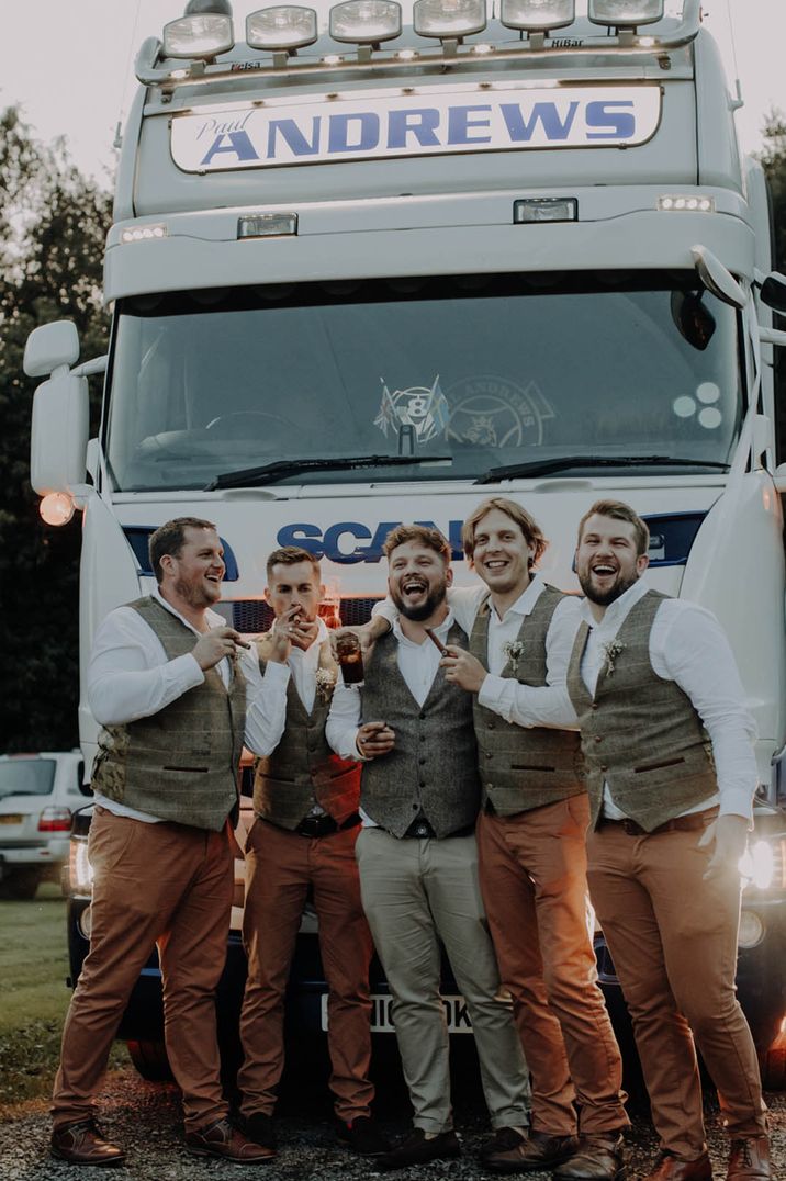 Groomsmen and groom smoking cigars and drinking beer for stag party with lorry wedding car