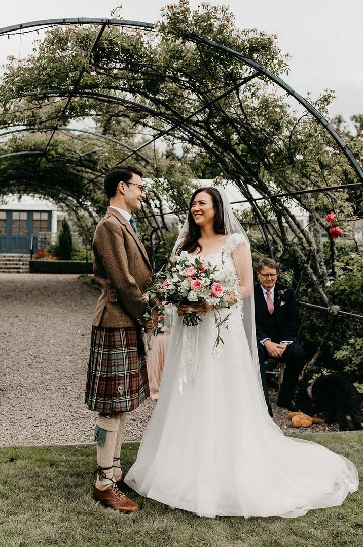 Groom in a brown suit with a kilt standing with the bride in a flower wedding dress with a pink and white bouquet 