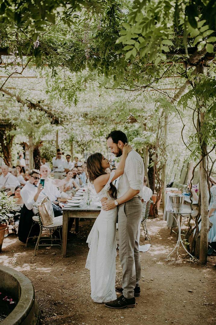 Bride in off the shoulder wedding dress and groom in white shirt and grey trousers embracing at Petersham Nurseries glasshouse wedding venue 