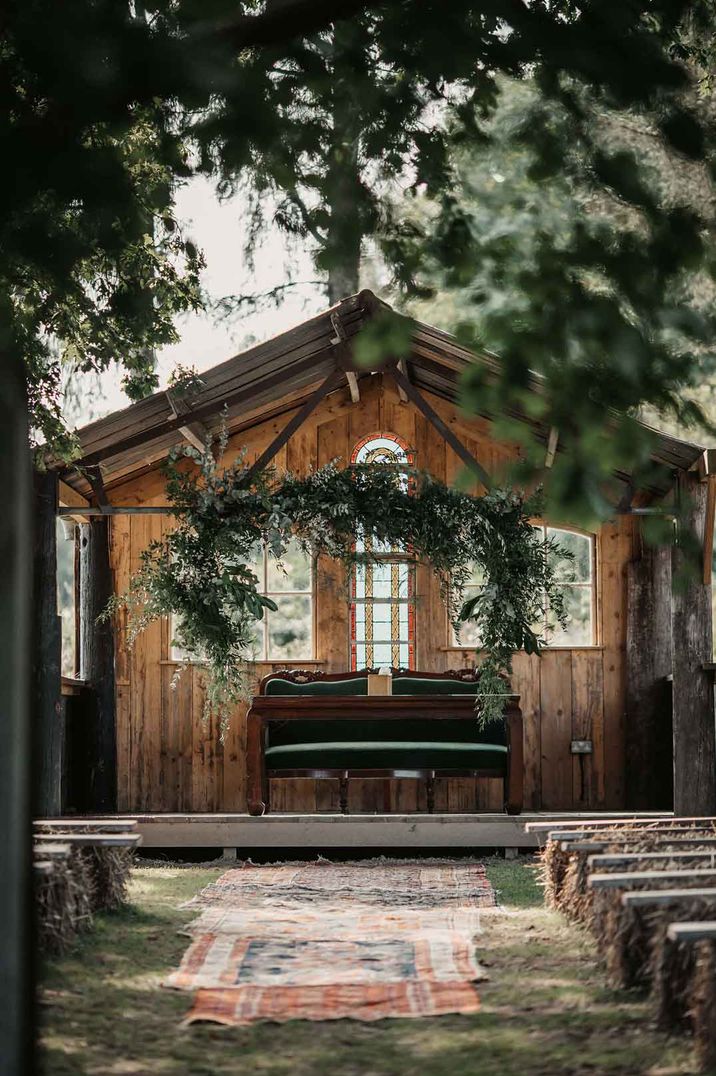 Boho rustic outdoor wedding alter with suspended foliage, forest green sofa, rugs gown down the alter and bench seats 