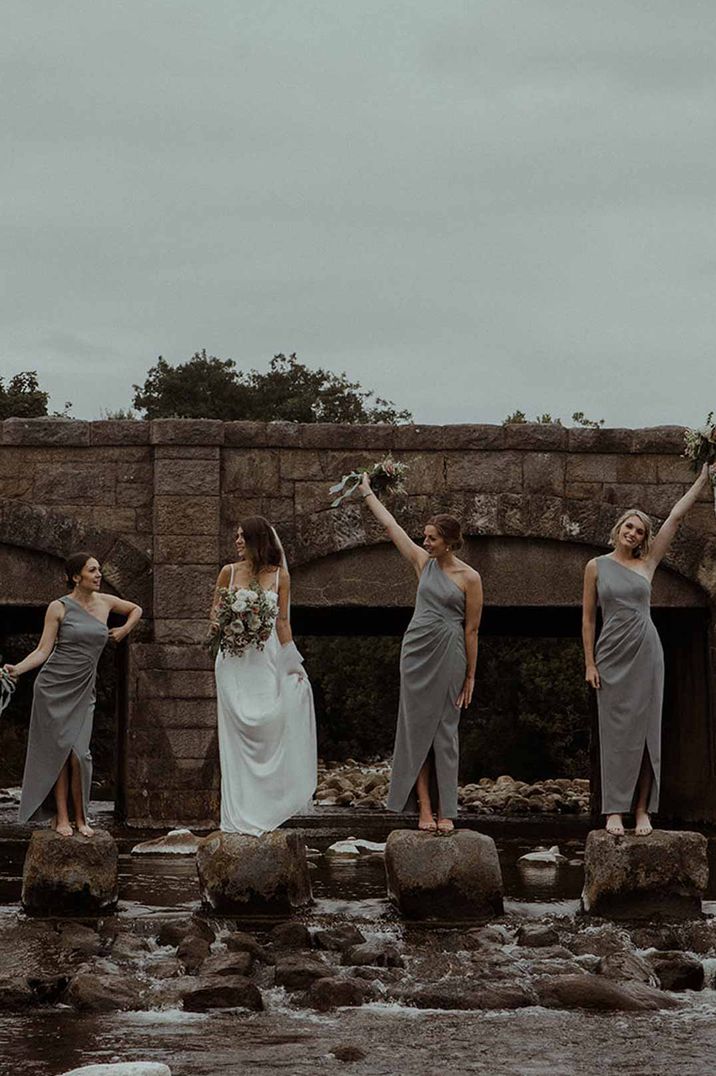 Bride in slip wedding dress standing on rocks with bridesmaids in marl grey one shoulder bridesmaid dresses outside The Dougarie Boathouse Scottish wedding venue