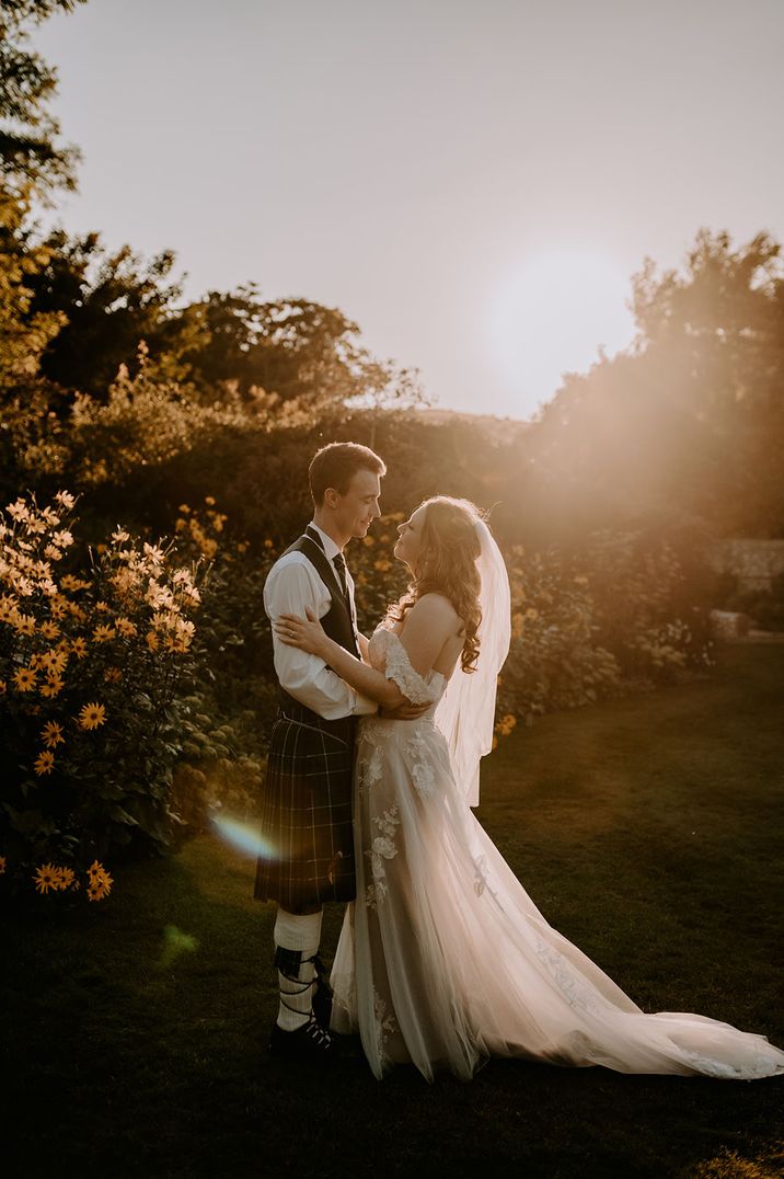 Groom in traditional Scottish kilt smiling at the bride in an off the shoulder lace wedding dress during the sunset 