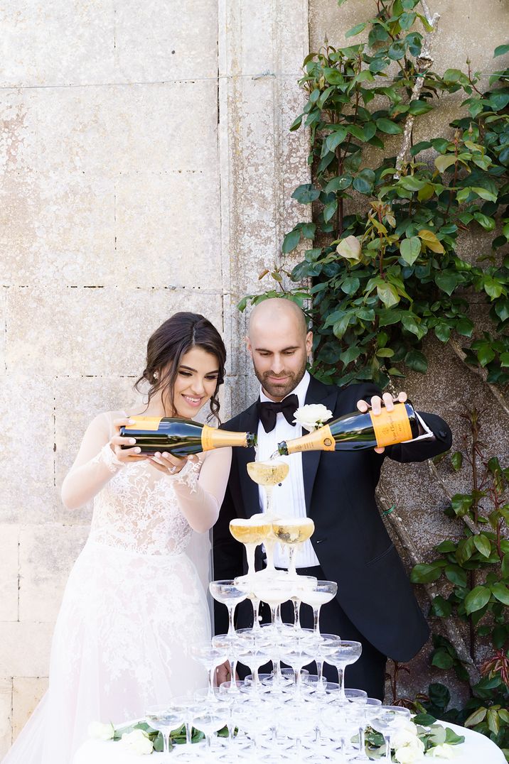 Bride in lace dress with sheer sleeves and groom in black tux pouring their champagne tower 