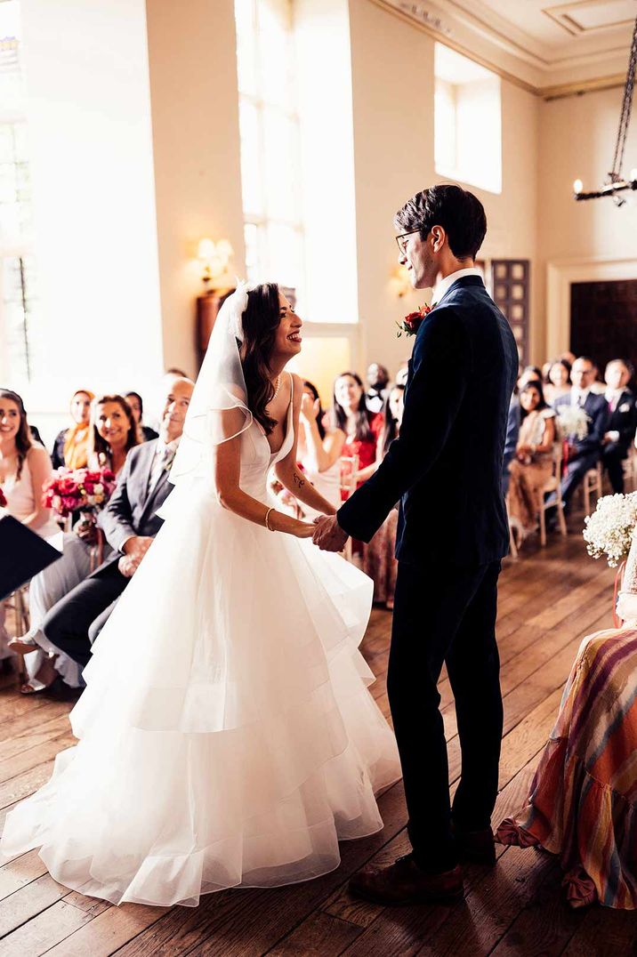 Bride in layered tulle wedding dress and veil stands holding hands with groom in dark suit and floral buttonhole during Elmore Court wedding ceremony