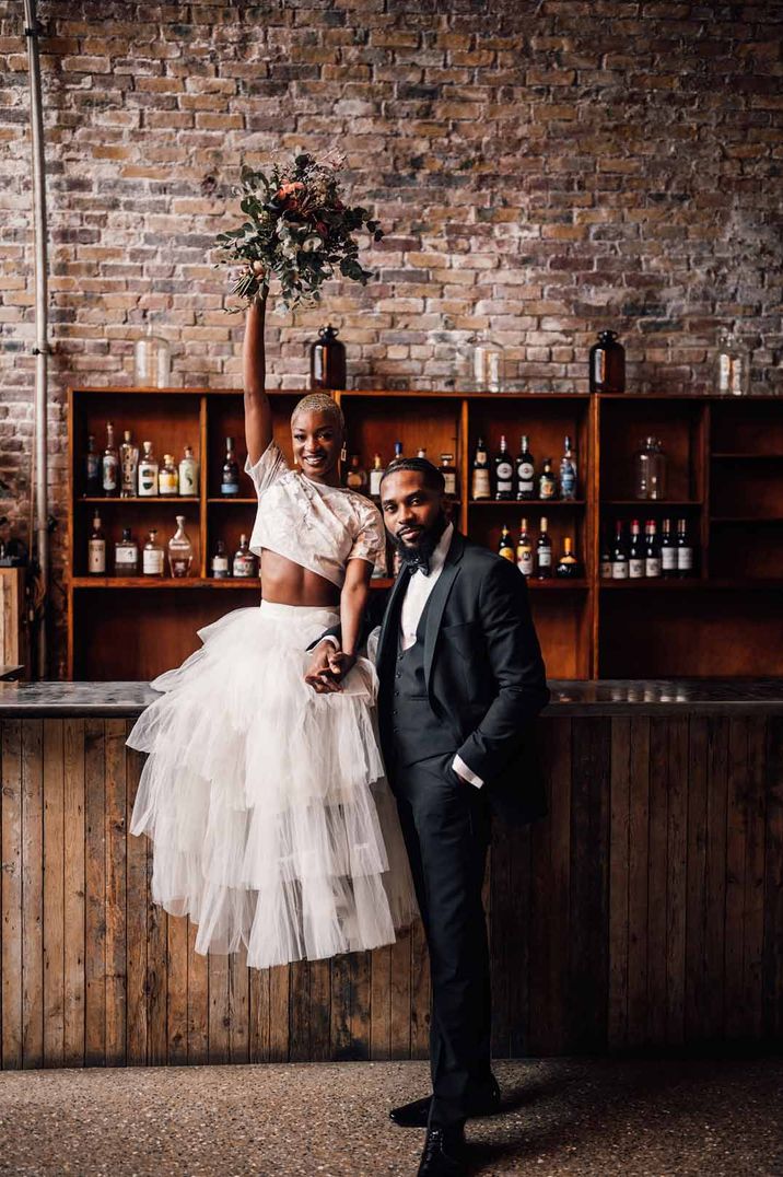 Bride in bridal two piece set with short sleeve lace crop top and layered tulle skirt sitting on the bar with groom in classic black tuxedo at 100 Barrington; exposed brick and rustic wedding bar 