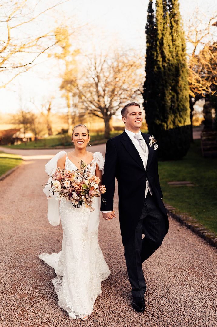 Bride in lace wedding dress with tulle wings walking along with the groom in a morning suit for traditional wedding 