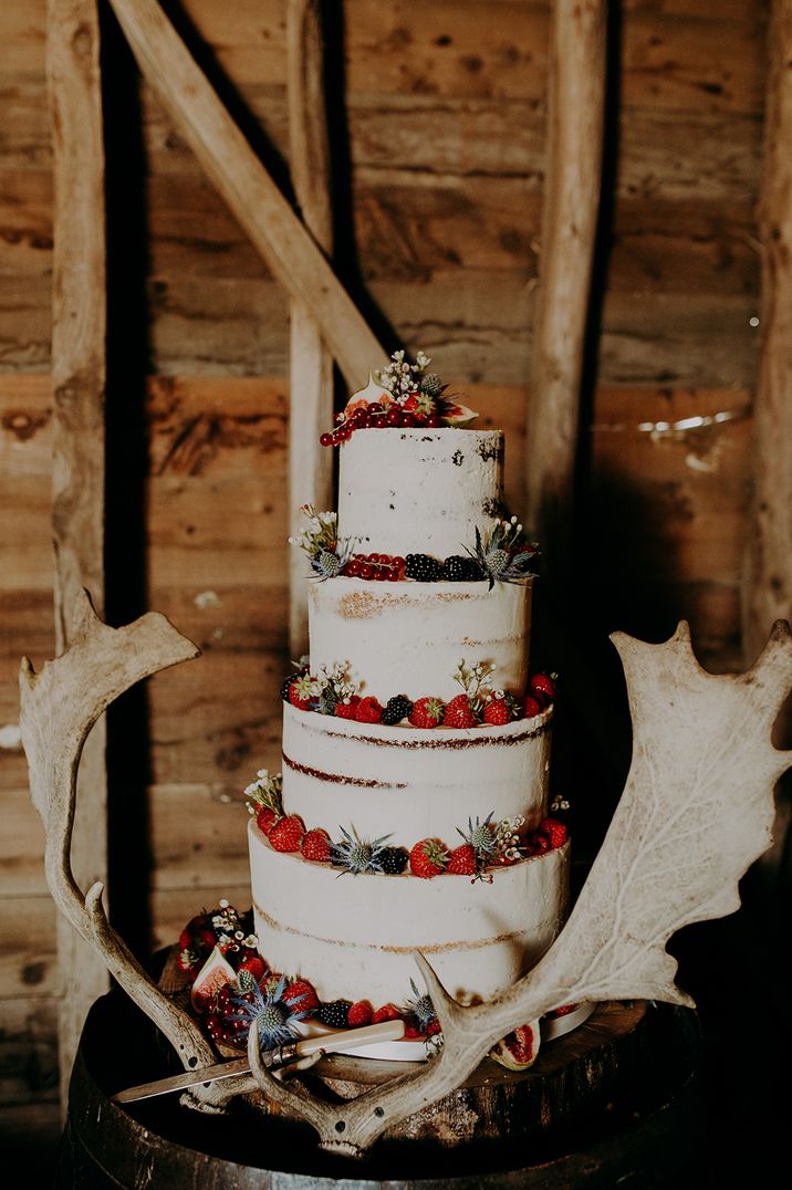 Semi naked wedding cake resting on a wooden barrel with fruit and antler wedding decor 