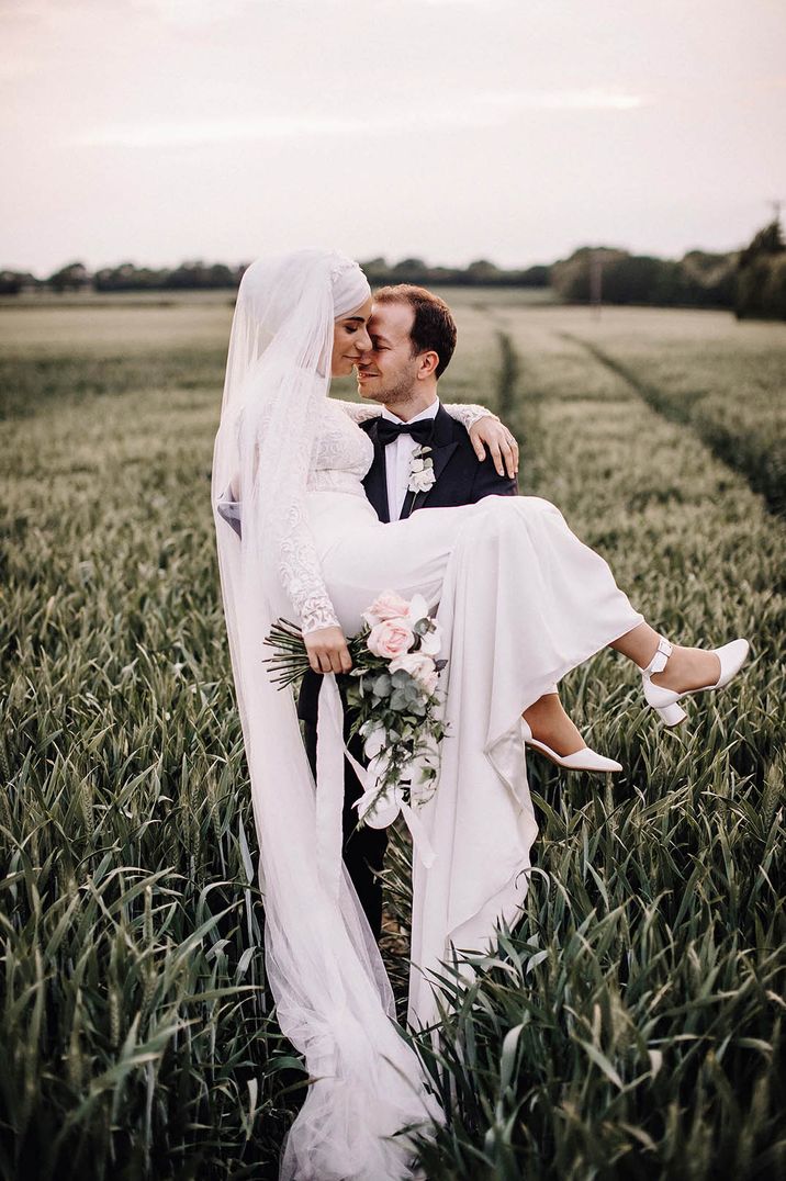 Muslim bride in white lace bridal outfit holding white rose and eucalyptus bouquet being held by groom in black tuxedo 