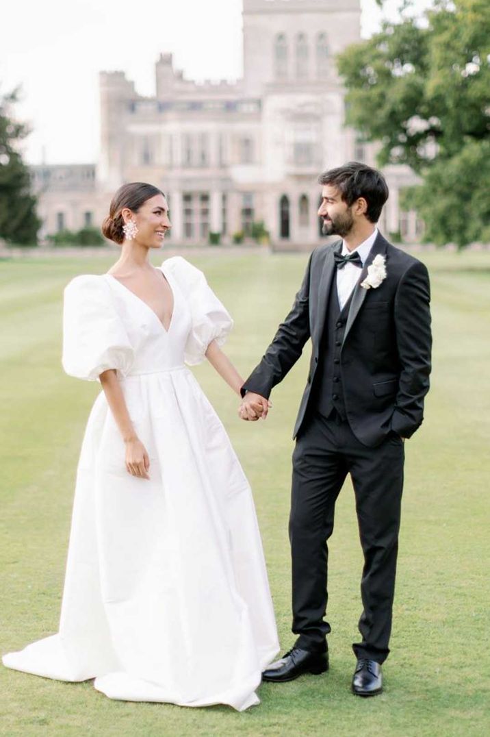 Bride in v neck puff sleeve wedding dress standing hand in hand with groom in classic black tuxedo at Ashridge House castle wedding venues