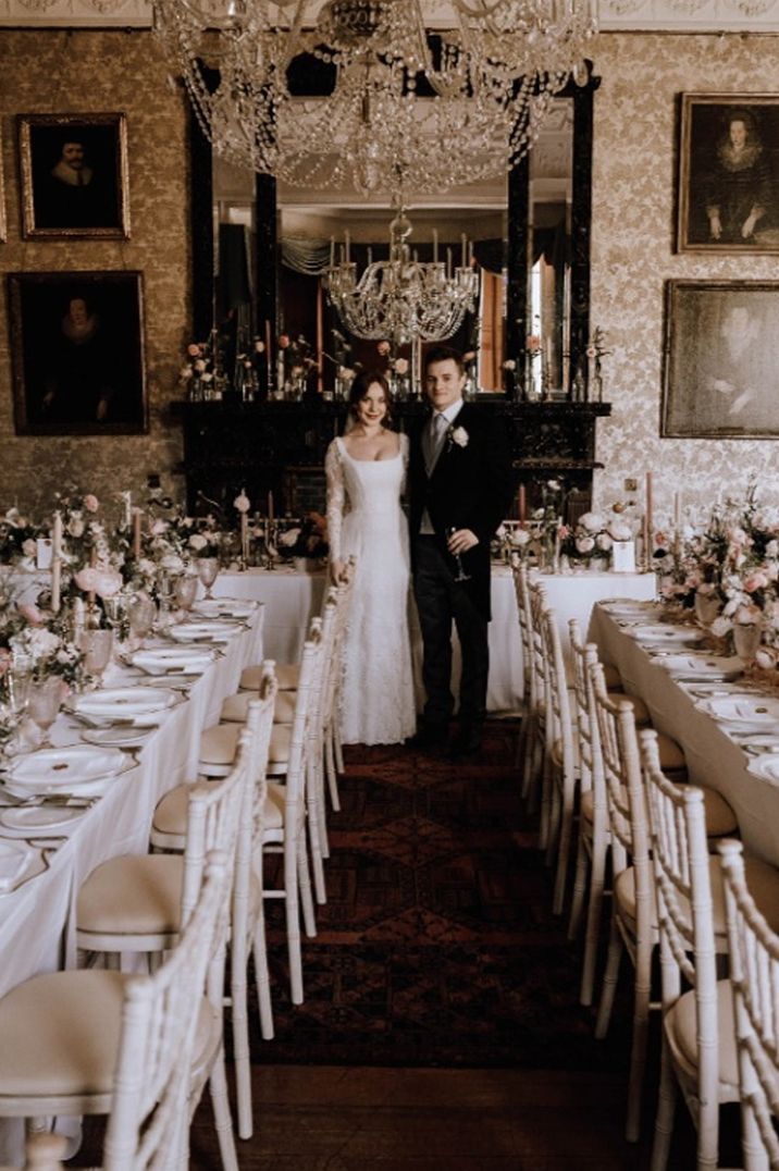 Bride in corset top style long sleeve wedding dress standing with groom in classic back grooms suit in reception room of Cambo House wedding venue with large chandeliers and classic wedding tablescapes 