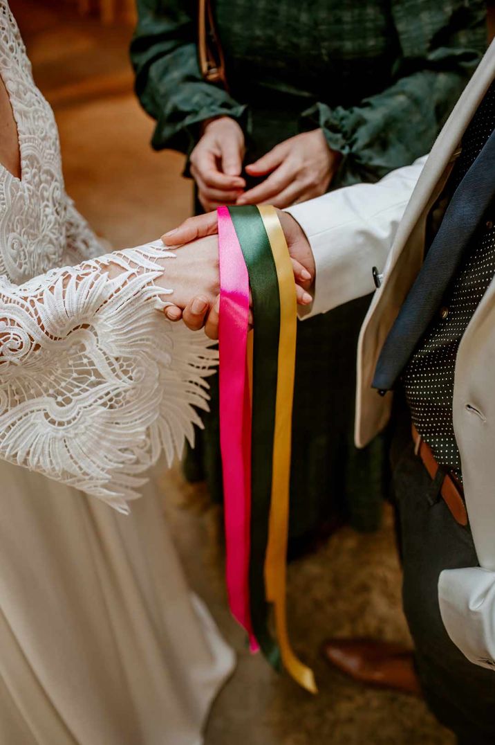 Bride in long sleeve lace boho wedding dress doing handfasting ceremony at wedding with groom in white suit 