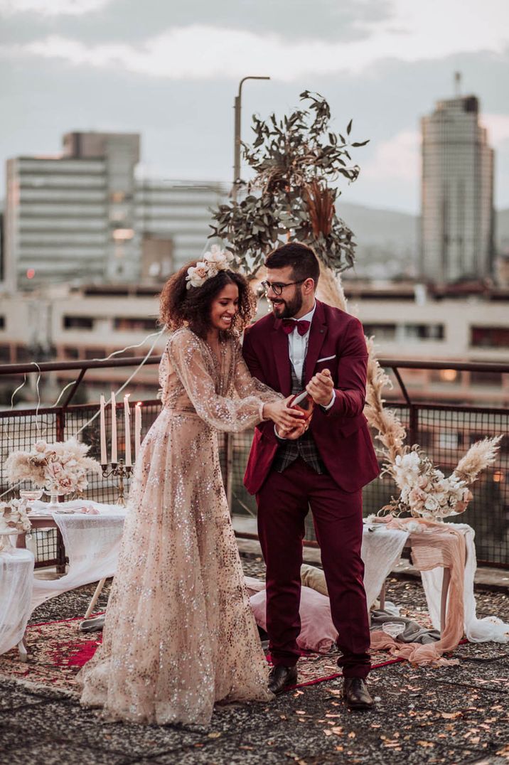 Bride in long balloon sleeve sparkly dress with groom in velvet suit with champagne bottle to celebrate 