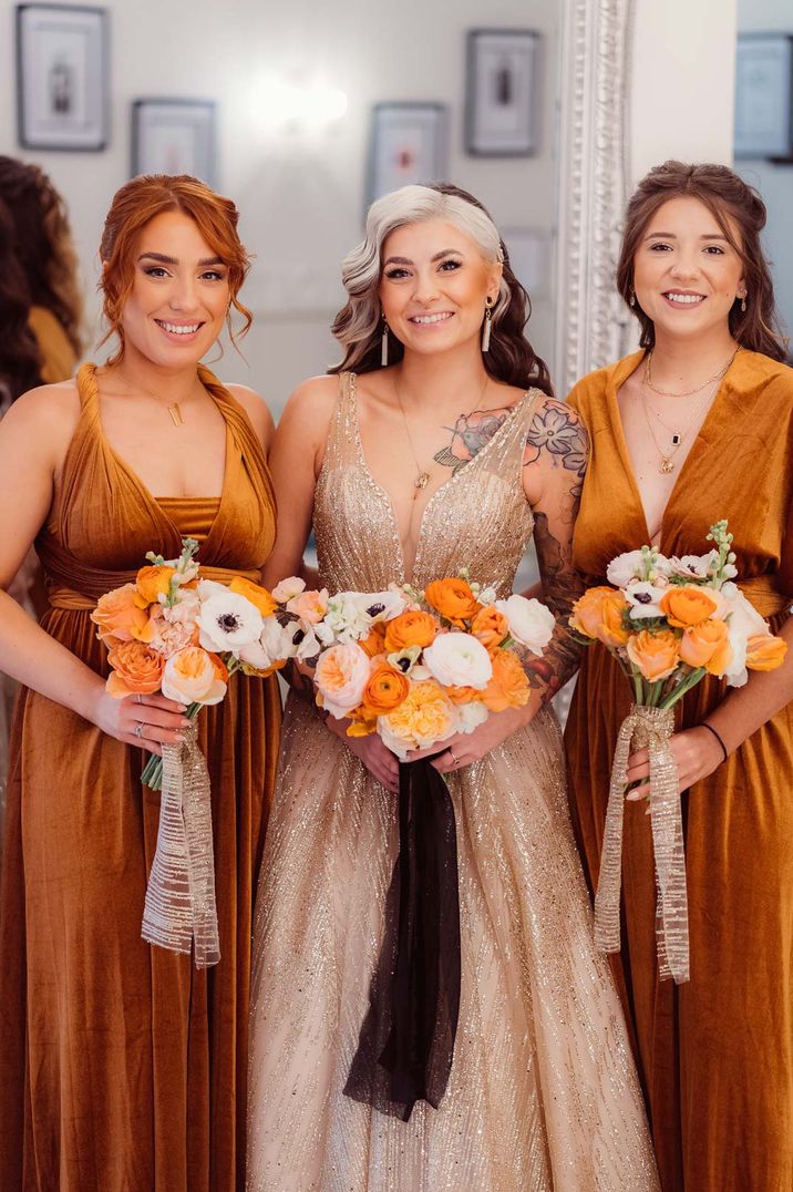 Bridal party wearing burnt orange velvet bridesmaid dresses holding orange and yellow wedding bouquets 