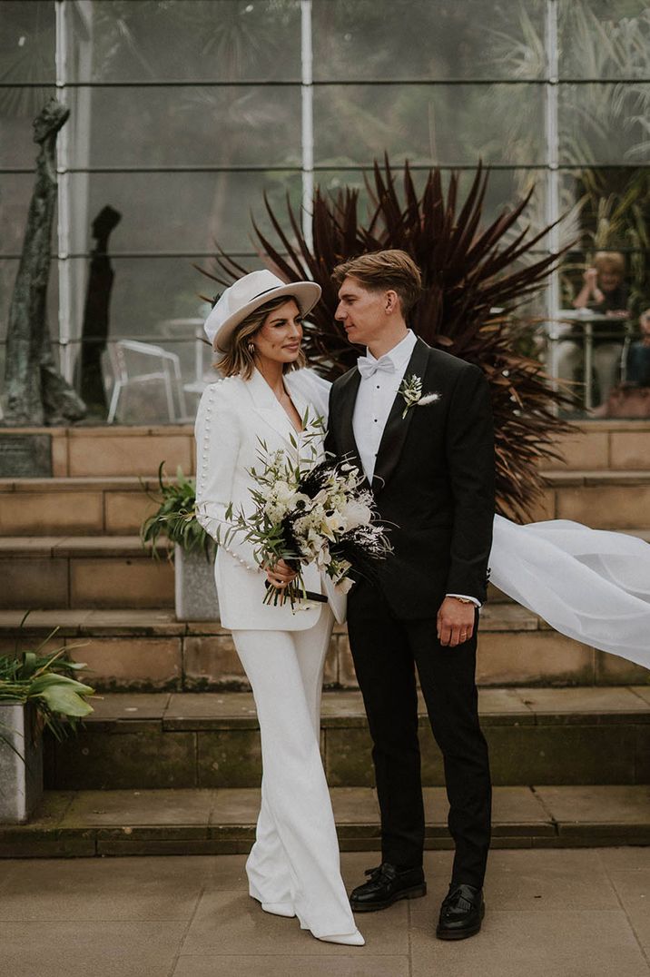 Groom in suit with grey bow tie smiling with bride in a Nadine Merabi pearl wedding suit with hat