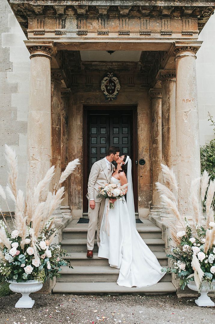 Groom in three piece cream wedding suit kissing the bride in a Jesus Peiro wedding dress at Elmore Court with pampas grass arrangements 