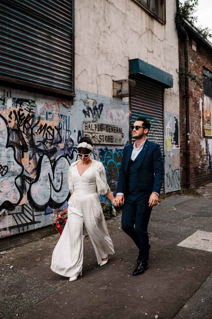 Bride in alternative bridal outfit - long sleeve v neck bridal jumpsuit with white wedding sunglasses and groom in classic grooms suit 