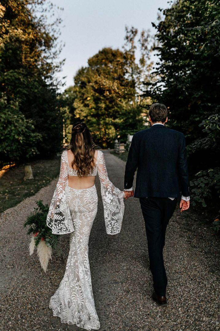 Bride in boho lace wedding dress with cutouts walking with the groom in a black suit for outdoor wedding 