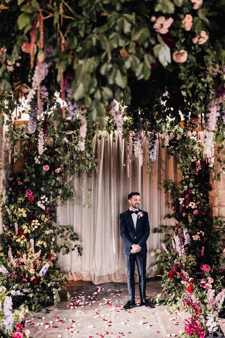 Groom in classic black tuxedo waiting in the reception room of Cripps Barn wedding venue with suspended foliage decor and large floral arches 