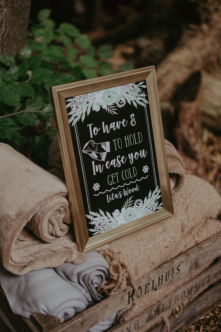 Wooden crate full of blankets with customised chalkboard signage at outdoor wedding 