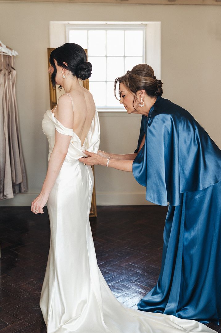 Mother of the bride in navy satin dress buttoning the bride up in her wedding dress