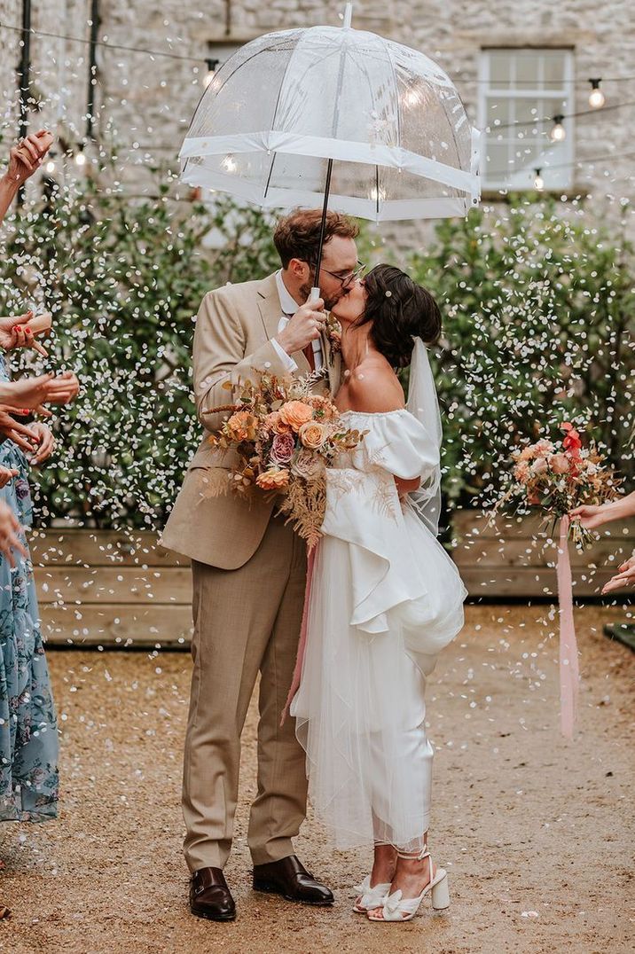 Bride and groom enjoy their confetti moment together as they kiss under a clear umbrella on rainy wedding day 