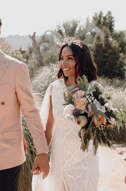 Groom in a pink double breasted jacket and floral bow tie holding hands with this bride in a lace boho wedding dress 