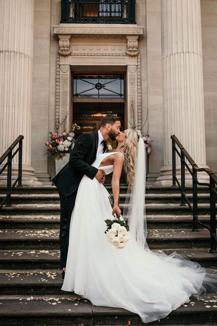 Groom in a tuxedo kissing his bride on the steps of Old Marylebone Town Hall in a princess wedding dress