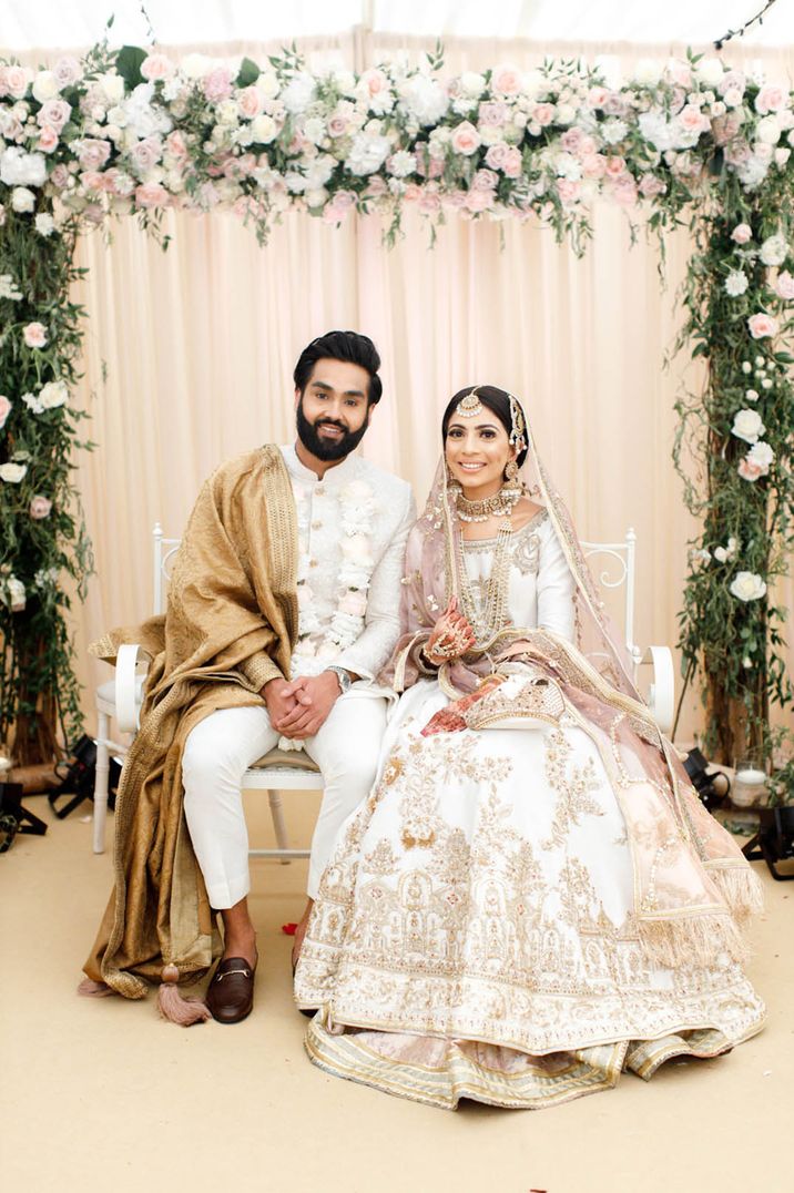 Pakistani Nikah wedding ceremony with the bride in a pink and white wedding outfit and the groom in white and gold with floral arch 