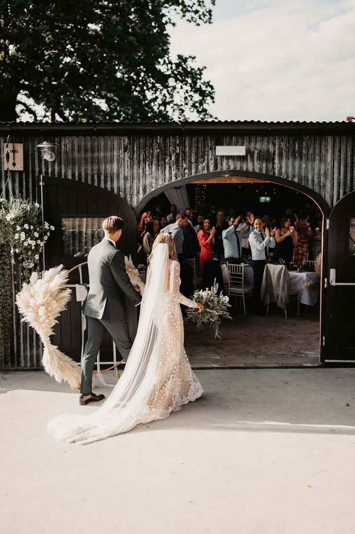 Bride in boho long sleeve sheer embellished wedding dress holding rose, peony, dried flower and eucalyptus bridal bouquet walking with groom into Oak Tree Barn wedding venue to guests cheering 