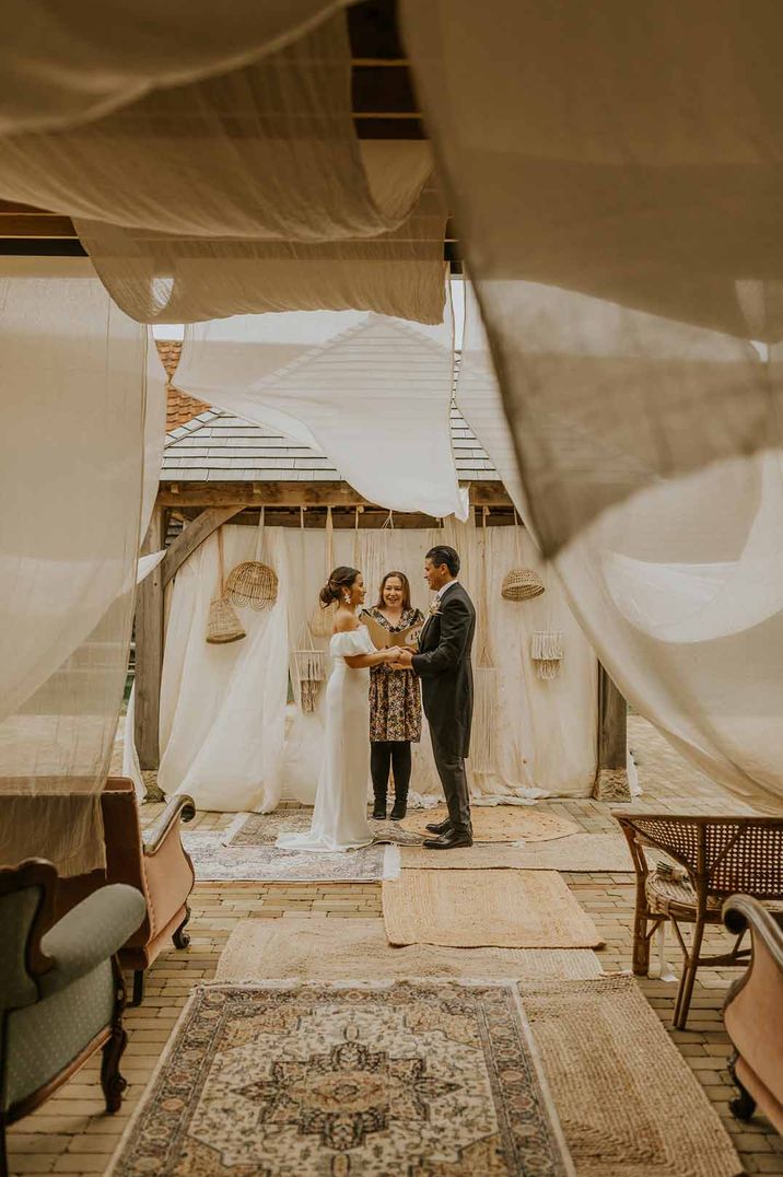 Bride in off shoulder wedding dress standing with groom in classic black tux at alter of The Canary Shed wedding venue with muted colour palette and wedding drapes 