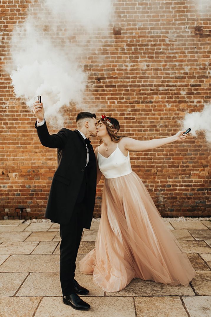 Bride in satin top wedding dress with pink tulle skirt with groom in black bow tie waving smoke bomb