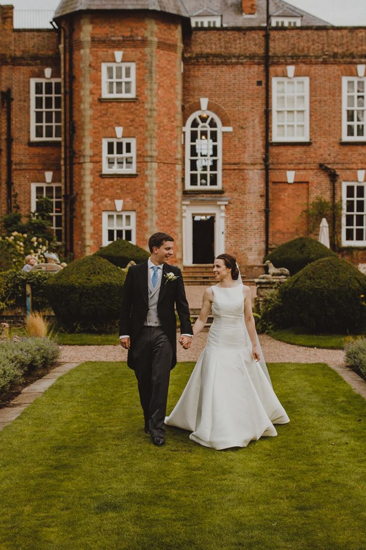 Bride in satin look sleeveless wedding dress and groom in dark suit walking round the grounds of Iscoyd Park