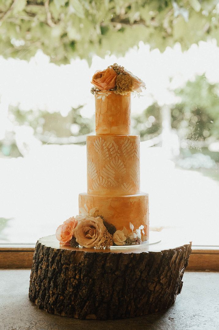 Three tier orange frosted wedding cake with pattern and flower decor on tree trunk stand for weddings under 40k