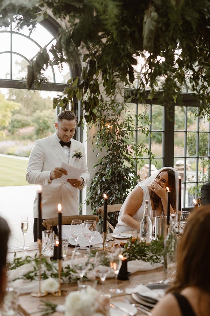 Groom in white tuxedo reading to bride in Made With Love wedding dress at rustic wedding tablescape with foliage and black tapered candles 