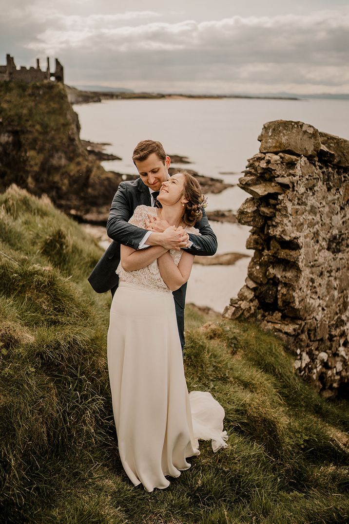 Groom embraces the bride from behind as they pose for couple portrait with ruins in the background 