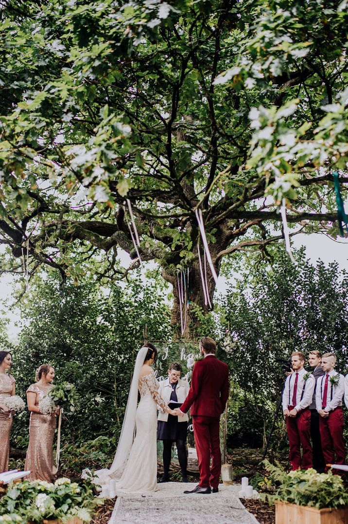 Outdoor woodland wedding ceremony with bride in a lace wedding dress and groom in a burgundy red wedding suit