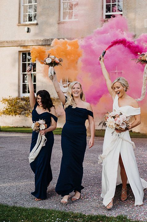 Bride in off the shoulder wedding dress and bridesmaids in navy blue dresses hold up orange and pink smoke bombs