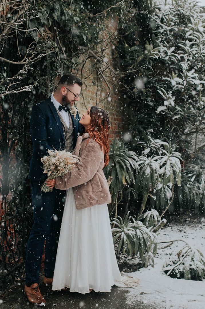 Bride in a brown fur coat stands looking up at the groom in a tartan waistcoat for their winter wedding